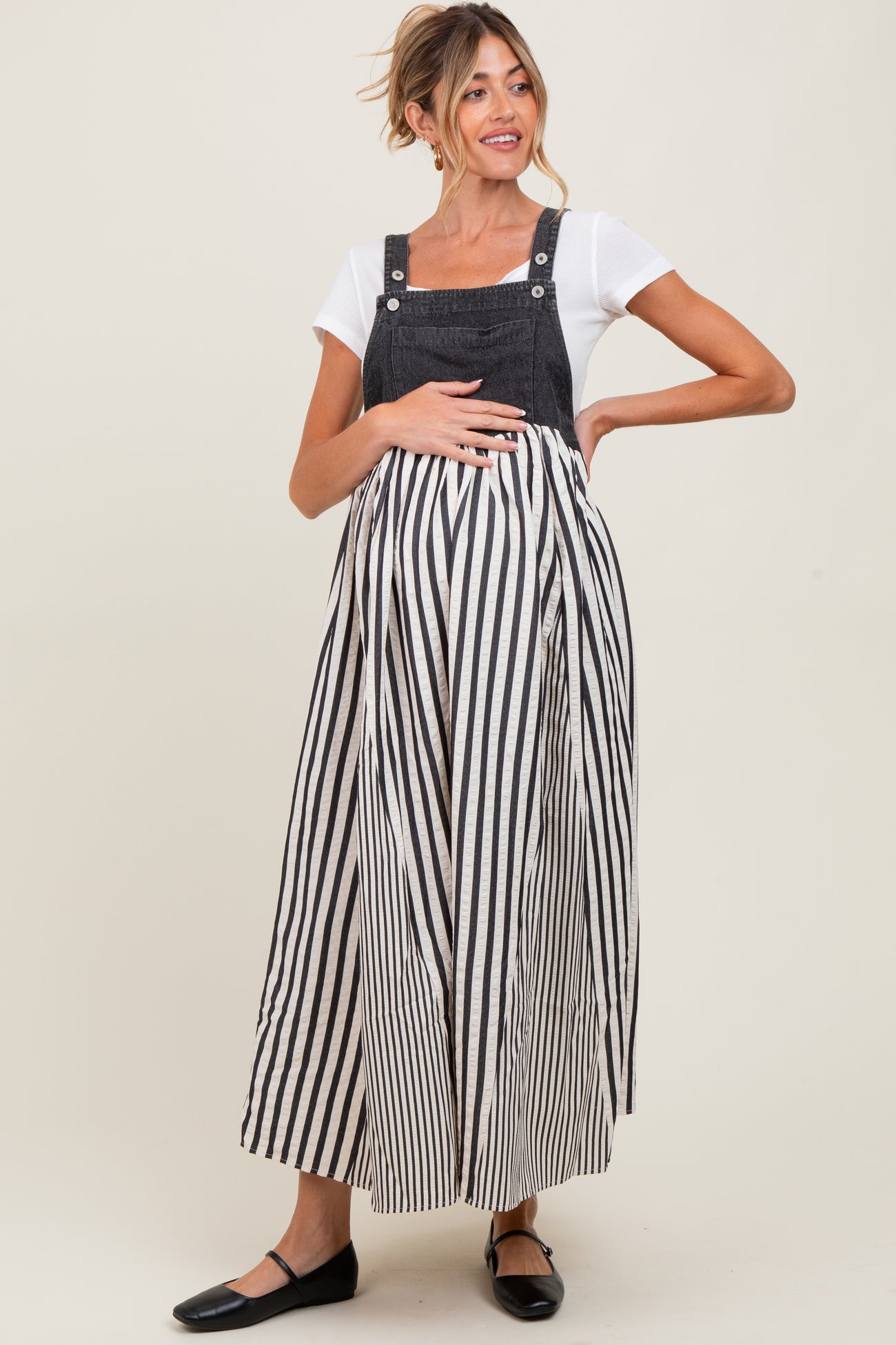 woman wearing a black and white striped maternity dress with denim overalls and holding her baby bump
