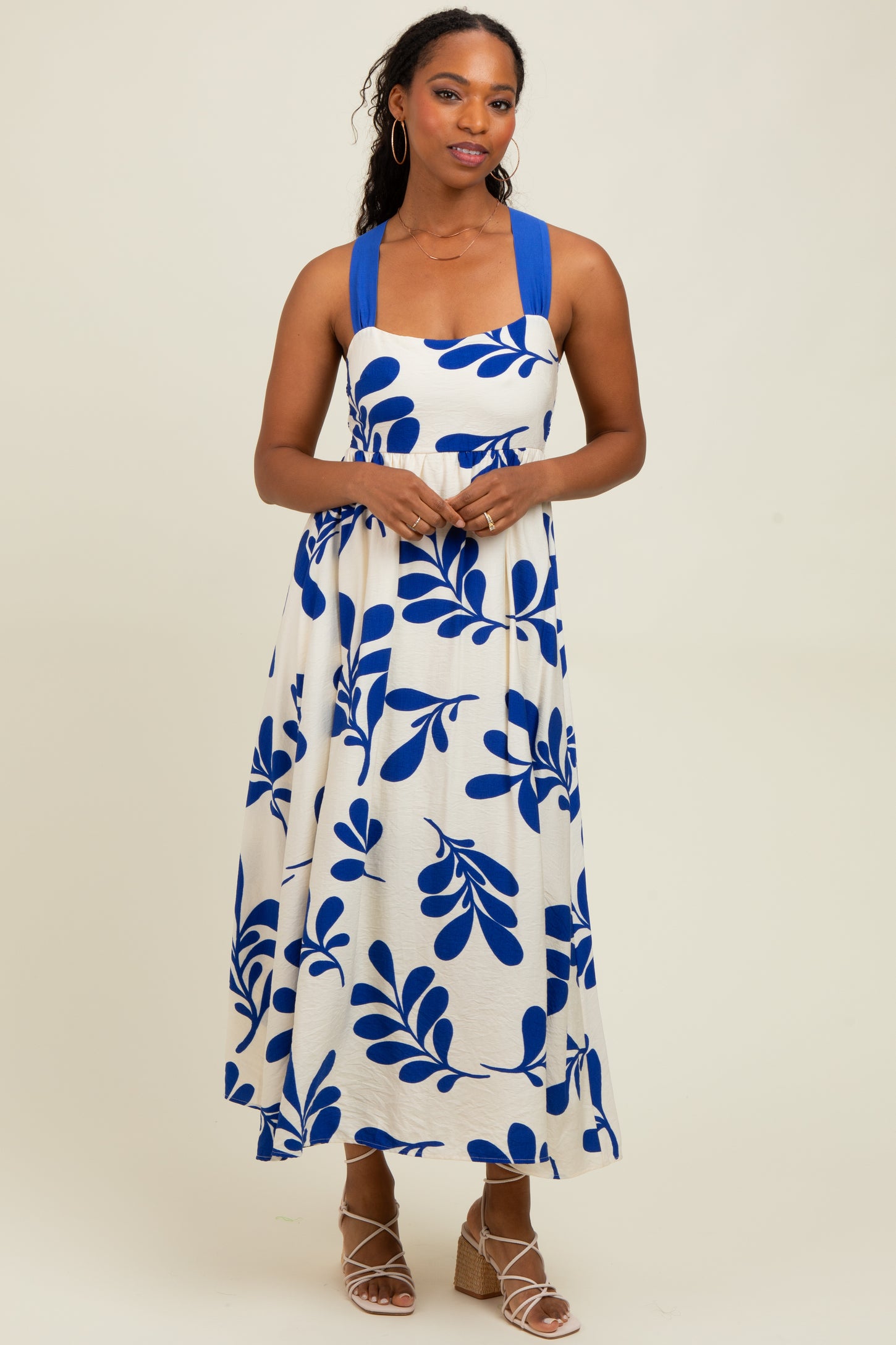 woman wearing a sleeveless white maxi dress with blue leaf print and blue straps, standing with hands clasped