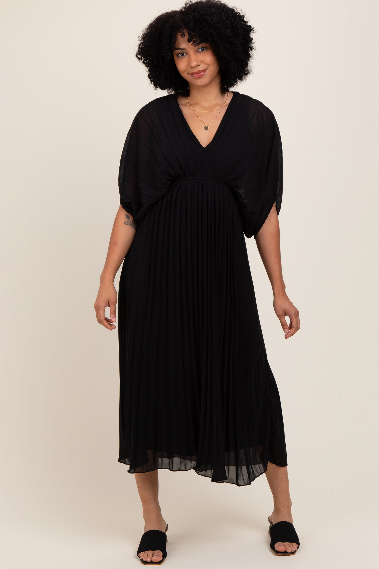 woman wearing a black pleated maxi dress with flutter sleeves and a v-neckline, posing with one hand on her hip