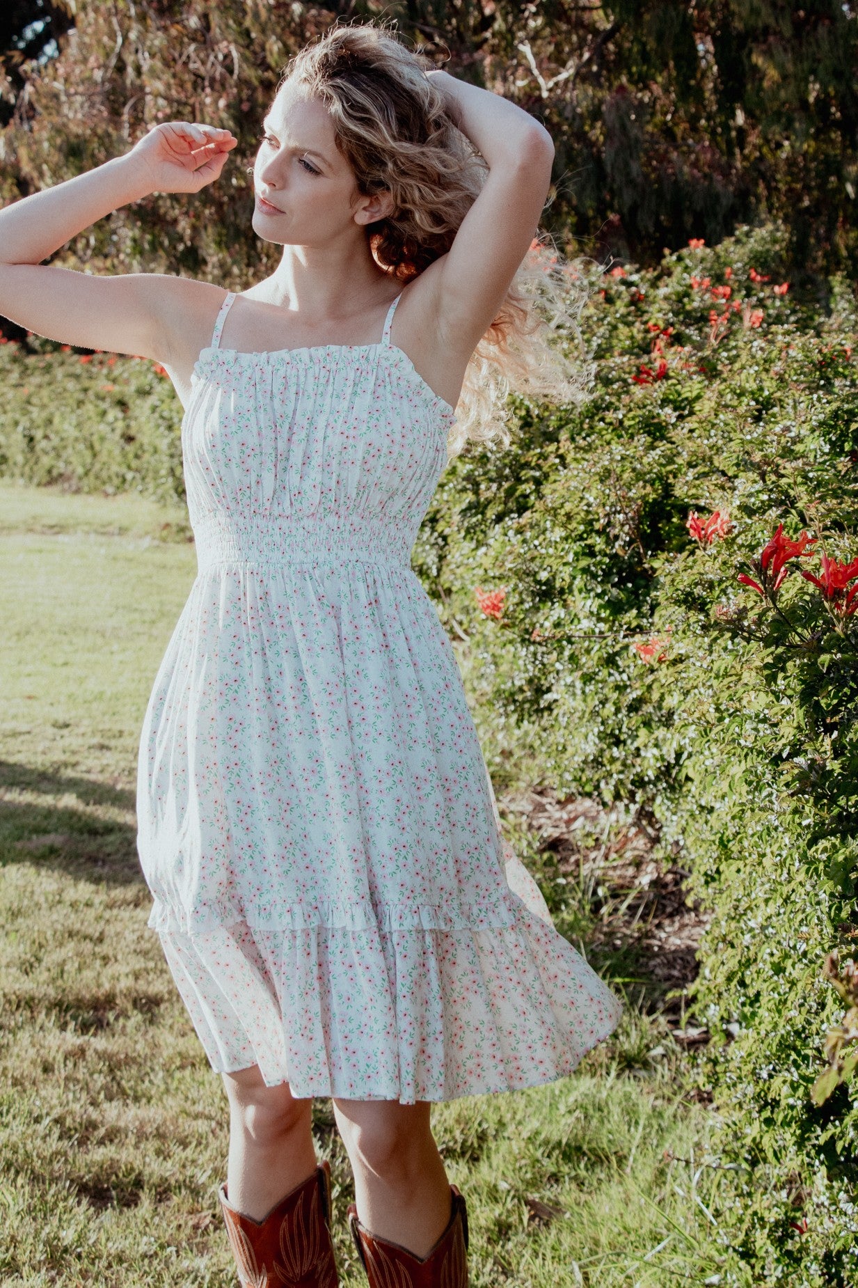 woman wearing a white floral sundress with a ruffled hem and spaghetti straps, posing outdoors among greenery and flowers