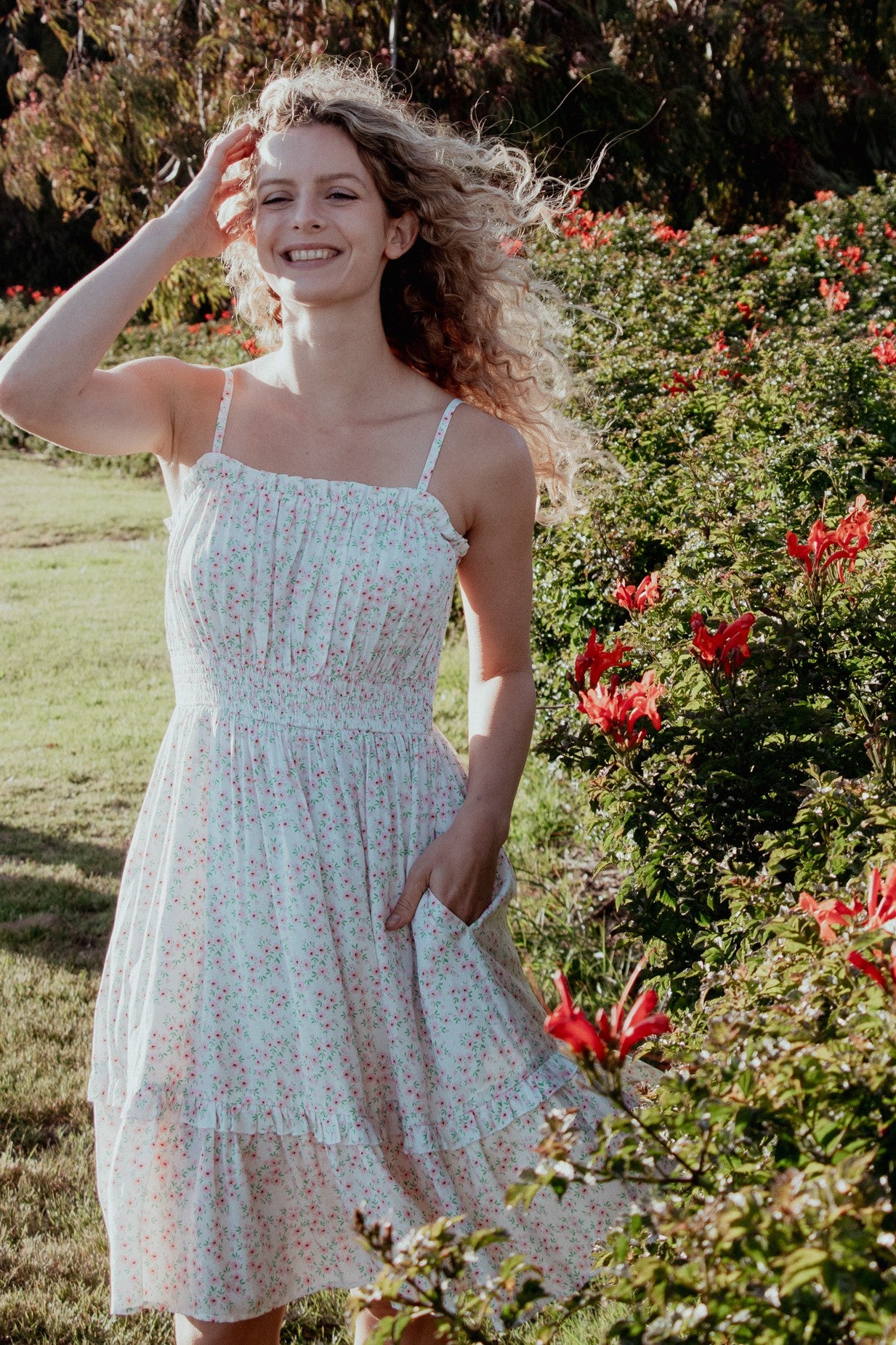 woman wearing a white floral sundress with thin straps and a tiered hem, posing in a garden with red flowers