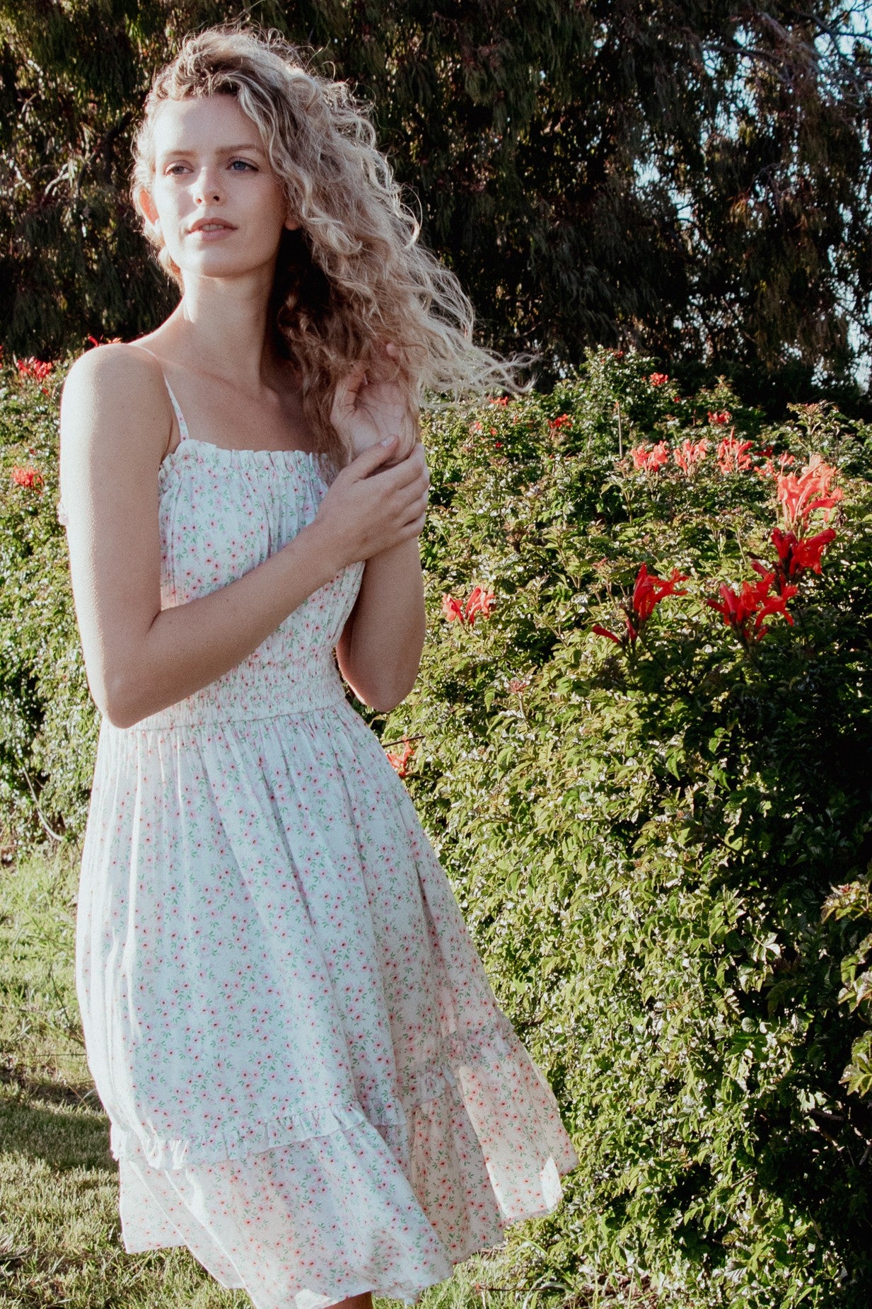 woman wearing a light pink floral sundress with smocked bodice, standing in a garden with blooming flowers