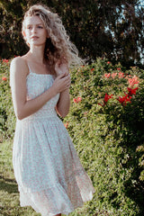 woman wearing a light pink floral sundress with smocked bodice, standing in a garden with blooming flowers