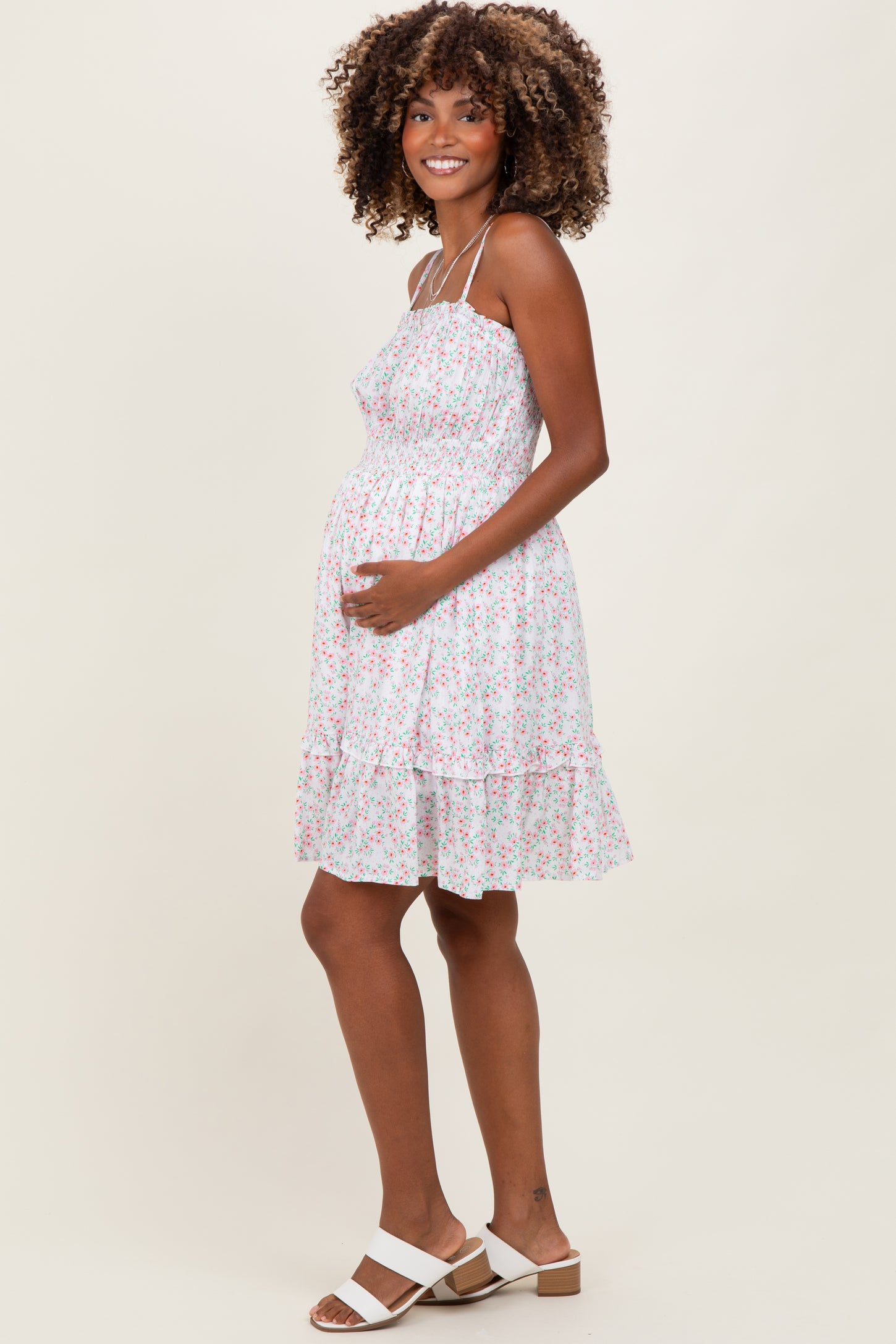 woman wearing a white floral maternity dress with spaghetti straps and a baby bump visible, smiling and holding her belly