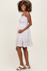 woman wearing a white floral maternity dress with spaghetti straps and a baby bump visible, smiling and holding her belly