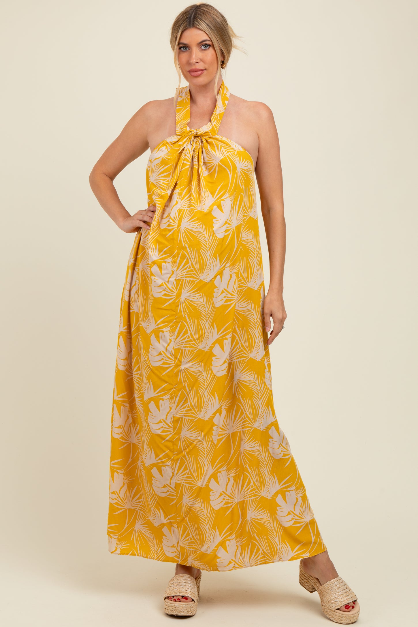 woman wearing a yellow halter neck maxi maternity dress with a tropical floral print and holding her baby bump