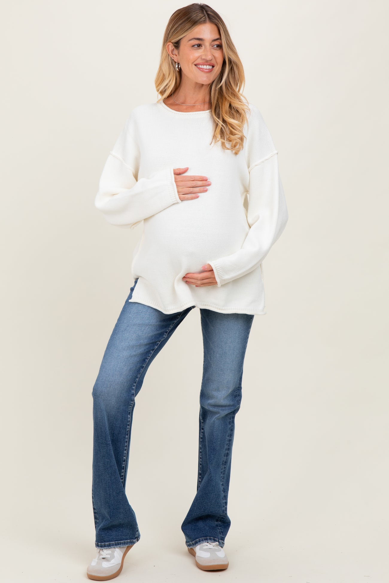 woman wearing a white oversized maternity sweater and blue flared jeans, holding her baby bump