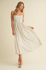 woman wearing a beige maxi dress with a square neckline and pleated skirt, standing elegantly