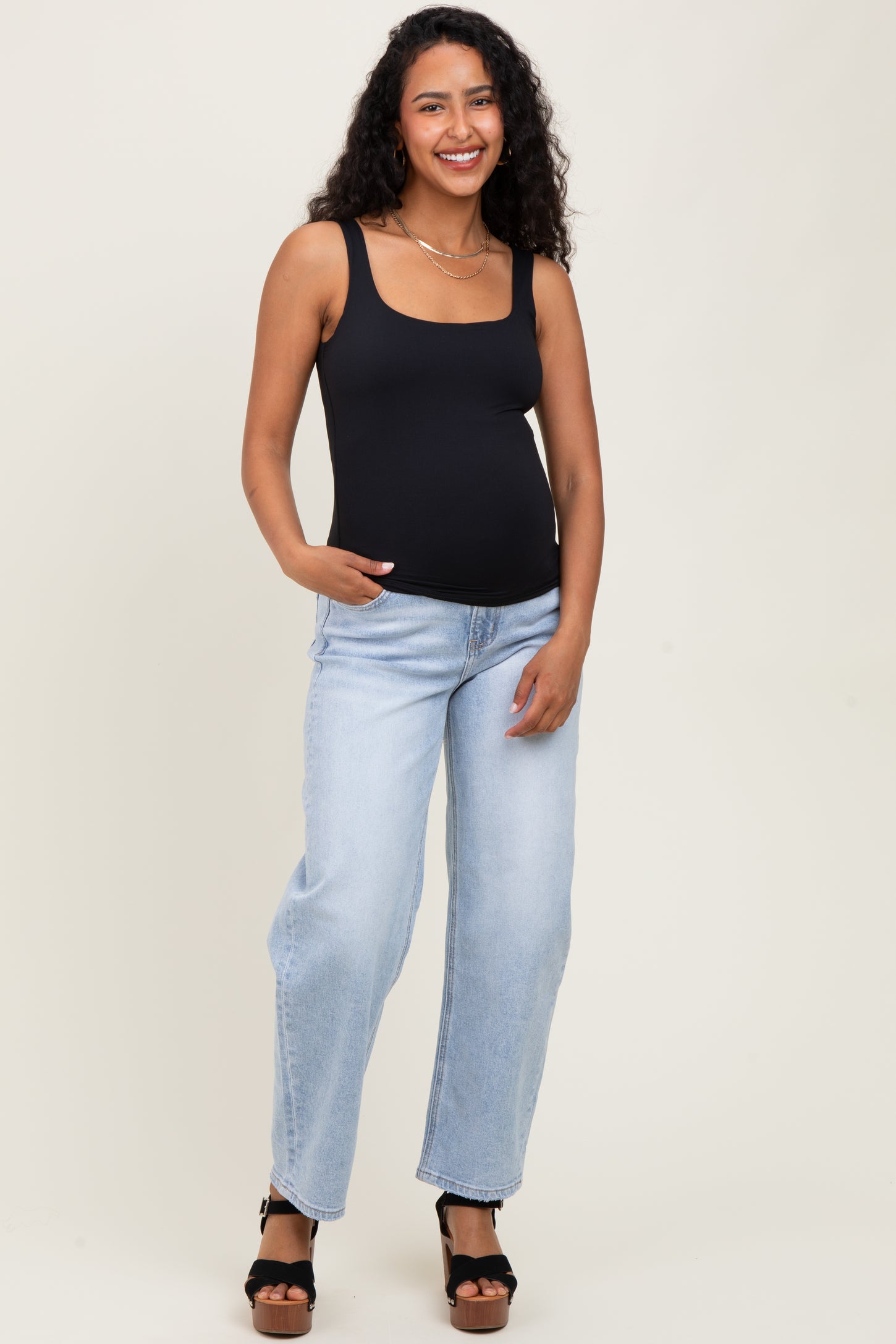 woman wearing a black maternity tank top and light blue distressed jeans, showing her baby bump while smiling