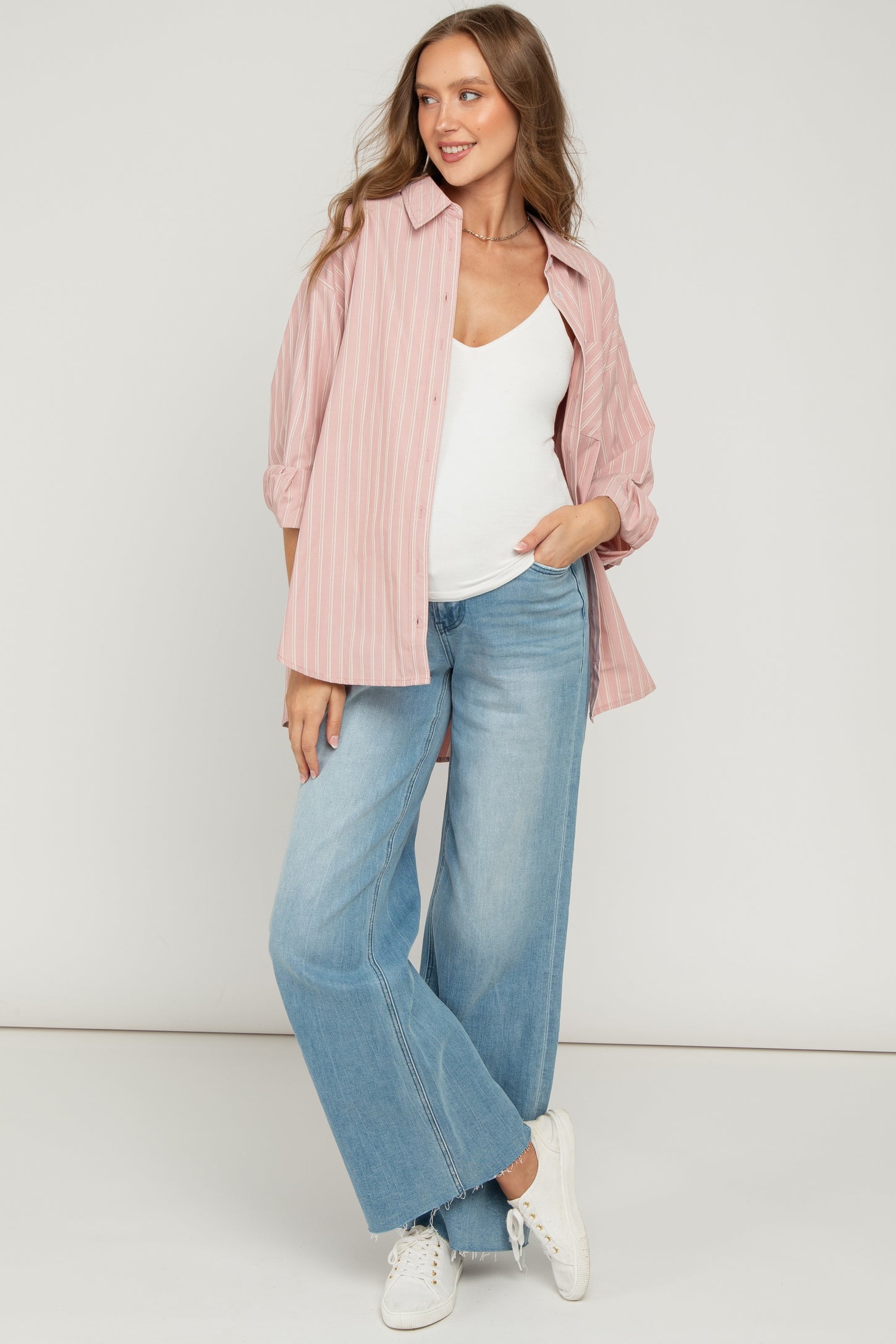 woman wearing a pink striped oversized button-up shirt and light blue wide-leg jeans, showing her baby bump