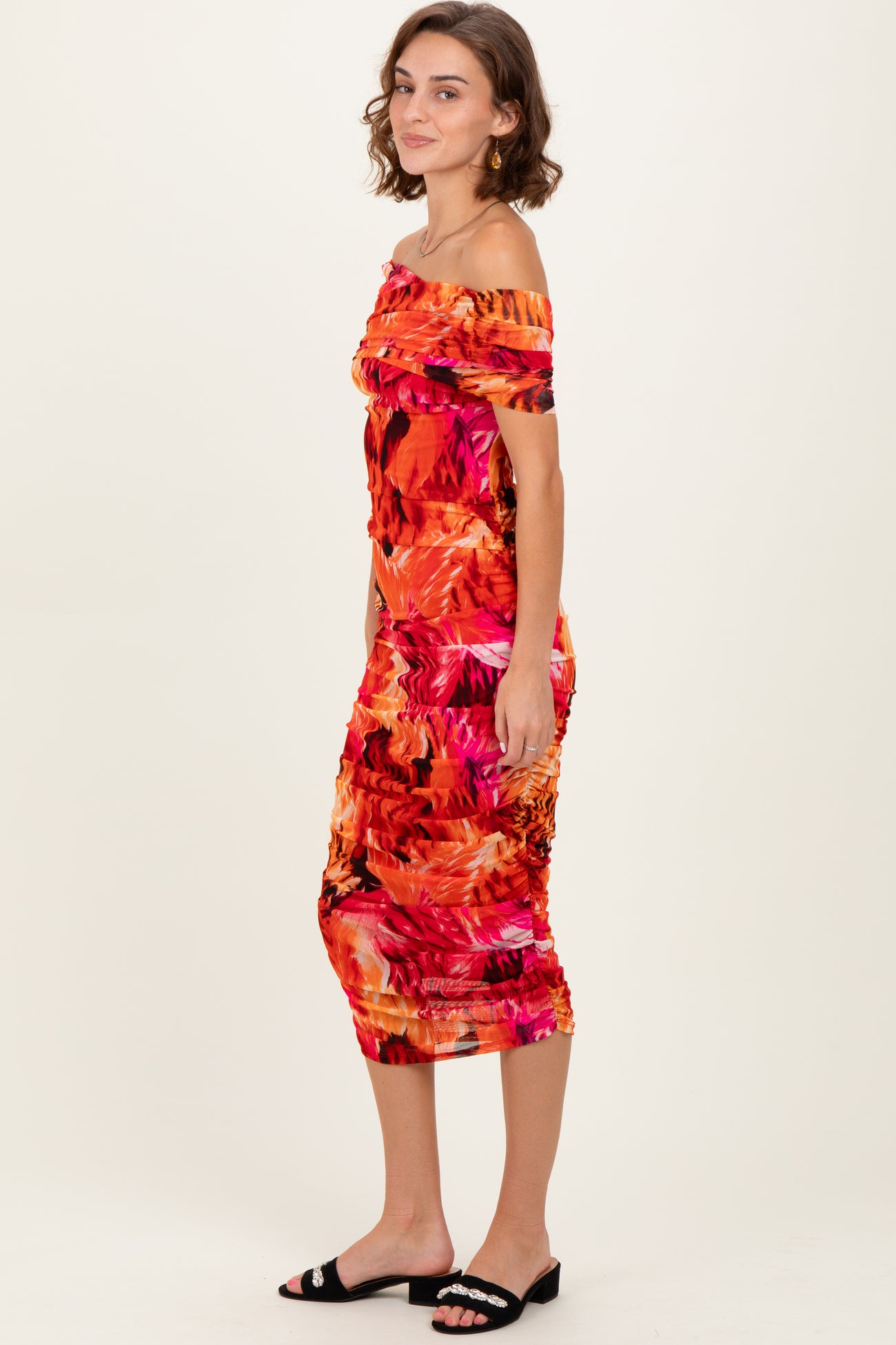 woman wearing a vibrant orange tropical print off-the-shoulder midi dress with ruching details and black slide sandals