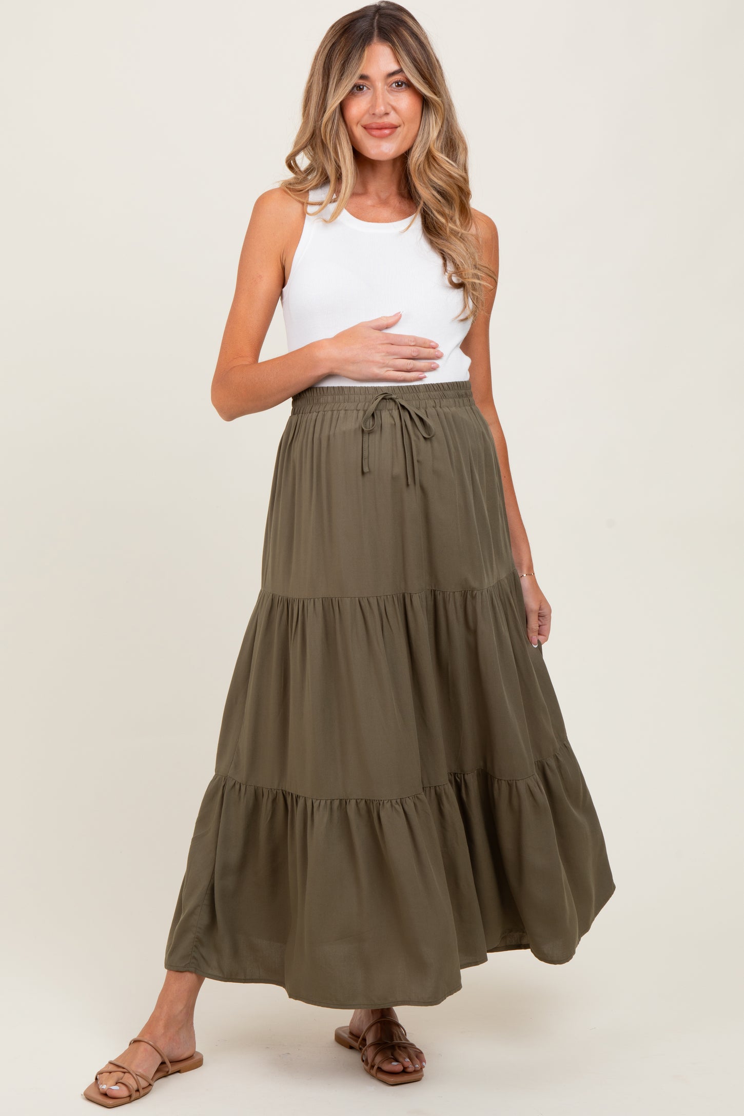 woman wearing a white tank top and an olive green tiered maternity skirt, holding her baby bump