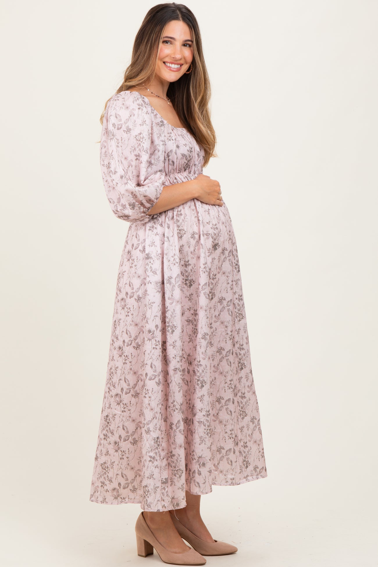 woman wearing a light pink floral maternity dress with a gathered waist and three-quarter sleeves, holding her baby bump