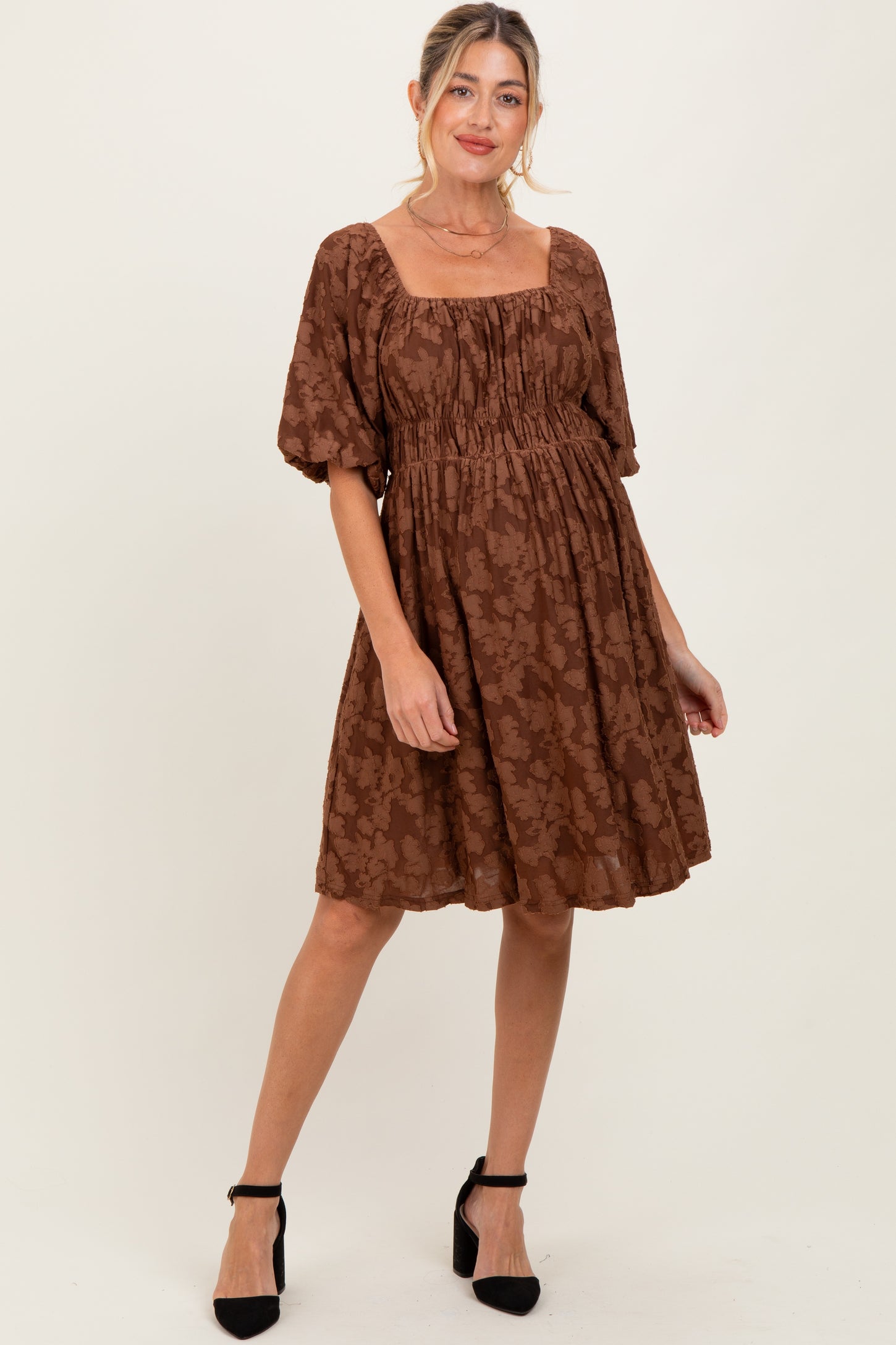 woman wearing a brown floral maternity dress with puff sleeves and a square neckline, standing and smiling
