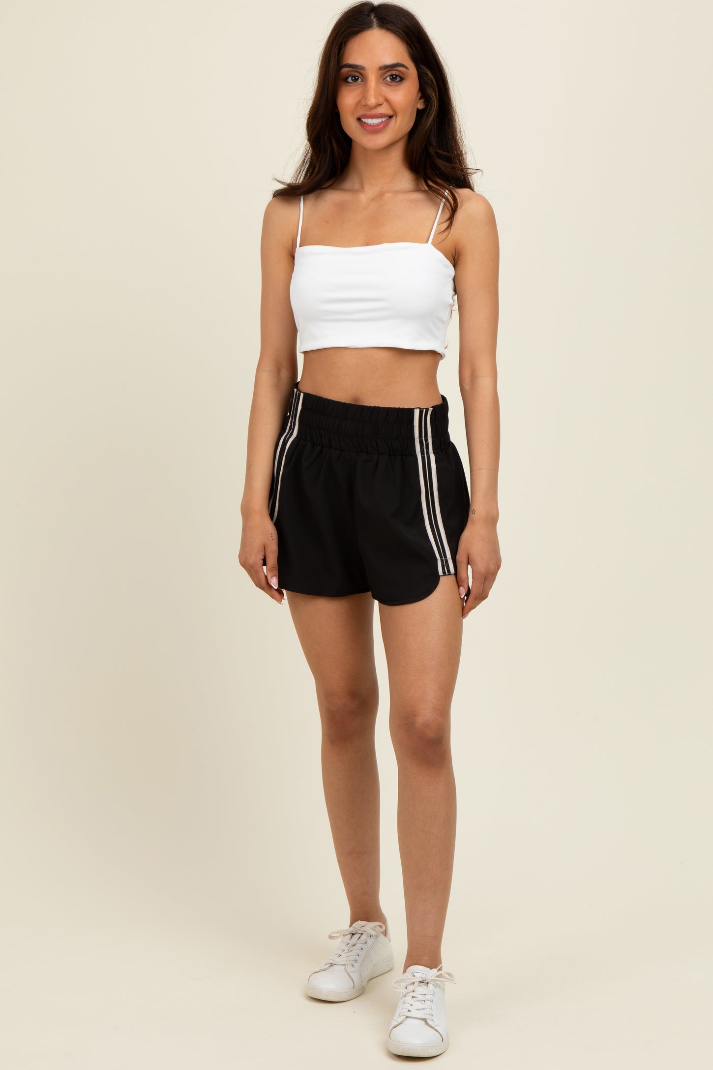 woman wearing a white cropped tank top and black athletic shorts with white side stripes, standing and smiling