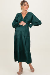 woman wearing a dark green maternity dress with a tiered design and v-neckline, holding her baby bump