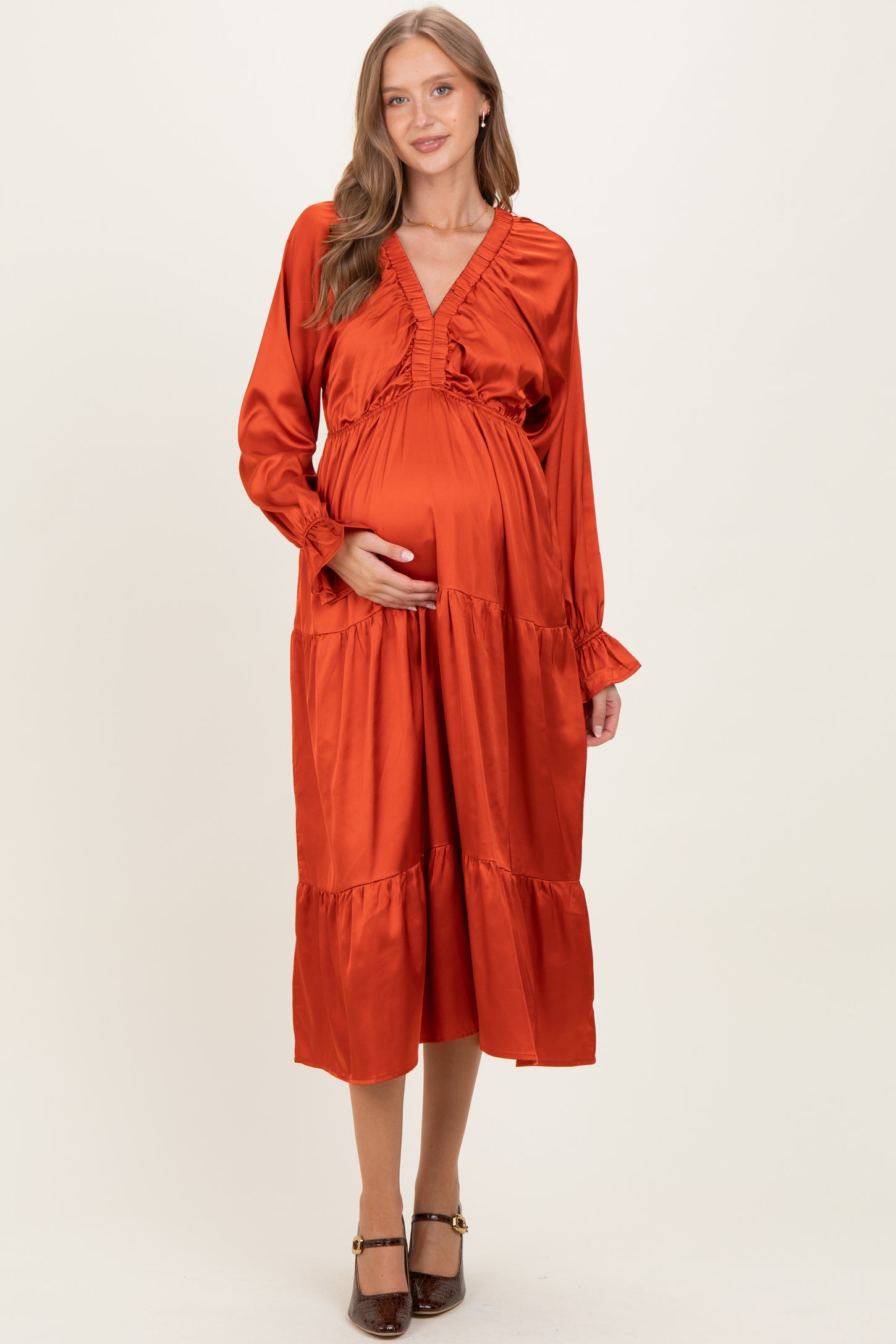 woman wearing an orange satin maternity dress with a v-neck and tiered skirt, holding her baby bump