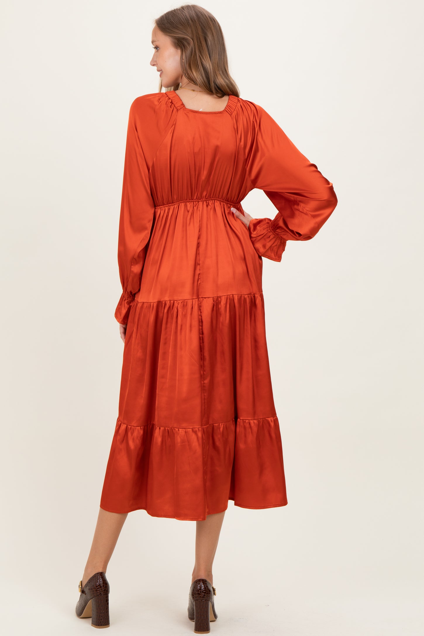 woman wearing a rust colored satin maternity dress with long sleeves and tiered skirt, showing a baby bump from the back
