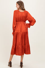 woman wearing a rust colored satin maternity dress with long sleeves and tiered skirt, showing a baby bump from the back