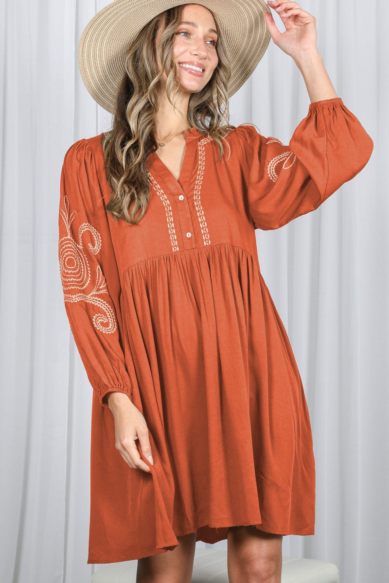 woman wearing an rust colored bohemian style dress with long sleeves and embroidered details, smiling while holding a hat