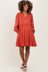 woman wearing a rust-colored maternity dress with long sleeves, decorative embroidery, and pockets, smiling at the camera