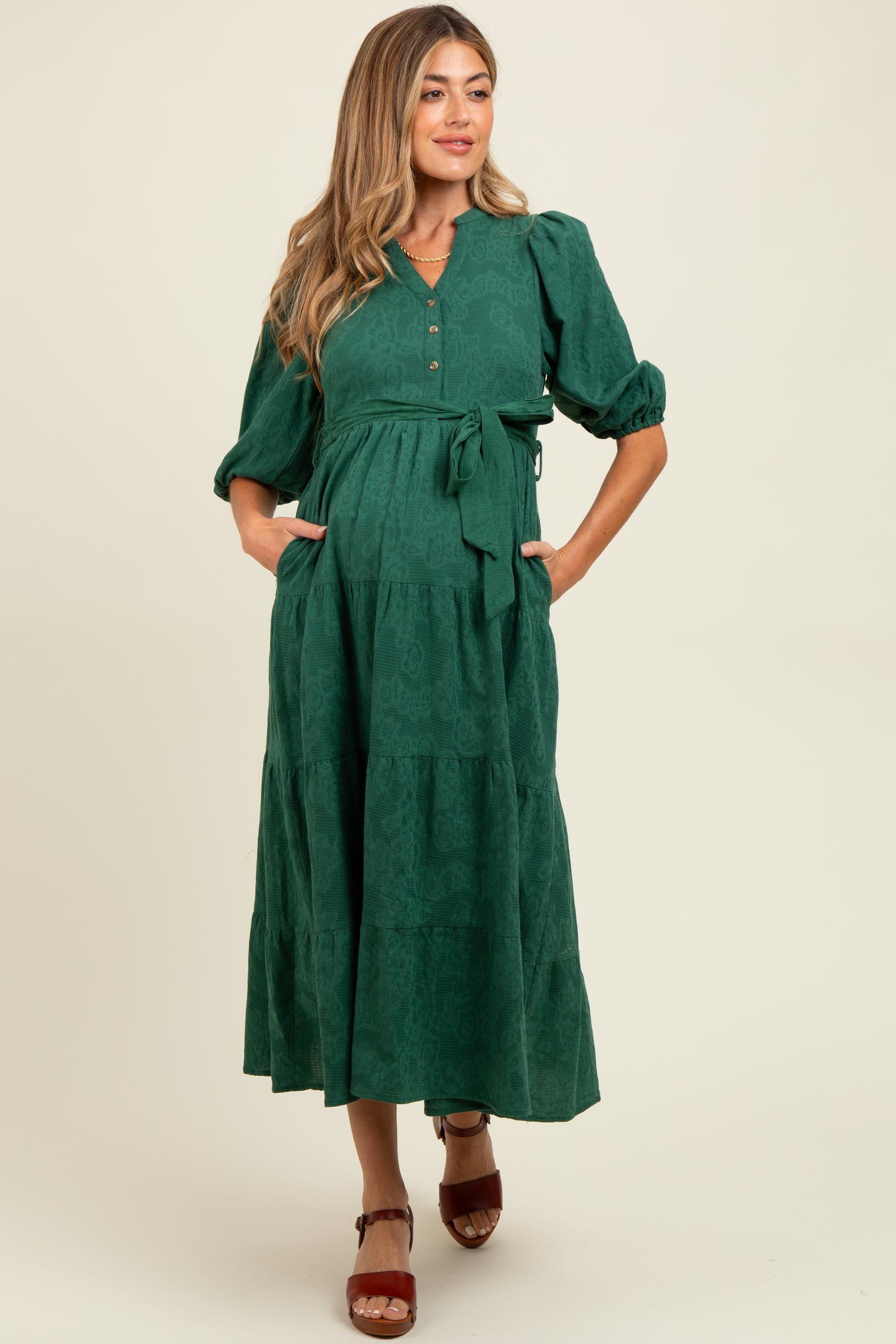 woman wearing a dark green maternity maxi dress with three-quarter sleeves, button details, and a tie belt, showing baby bump