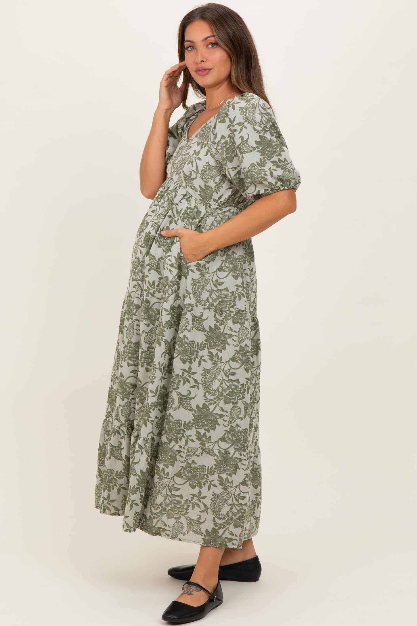 woman wearing a green floral print maternity dress with pockets and a gathered waist, showcasing her baby bump
