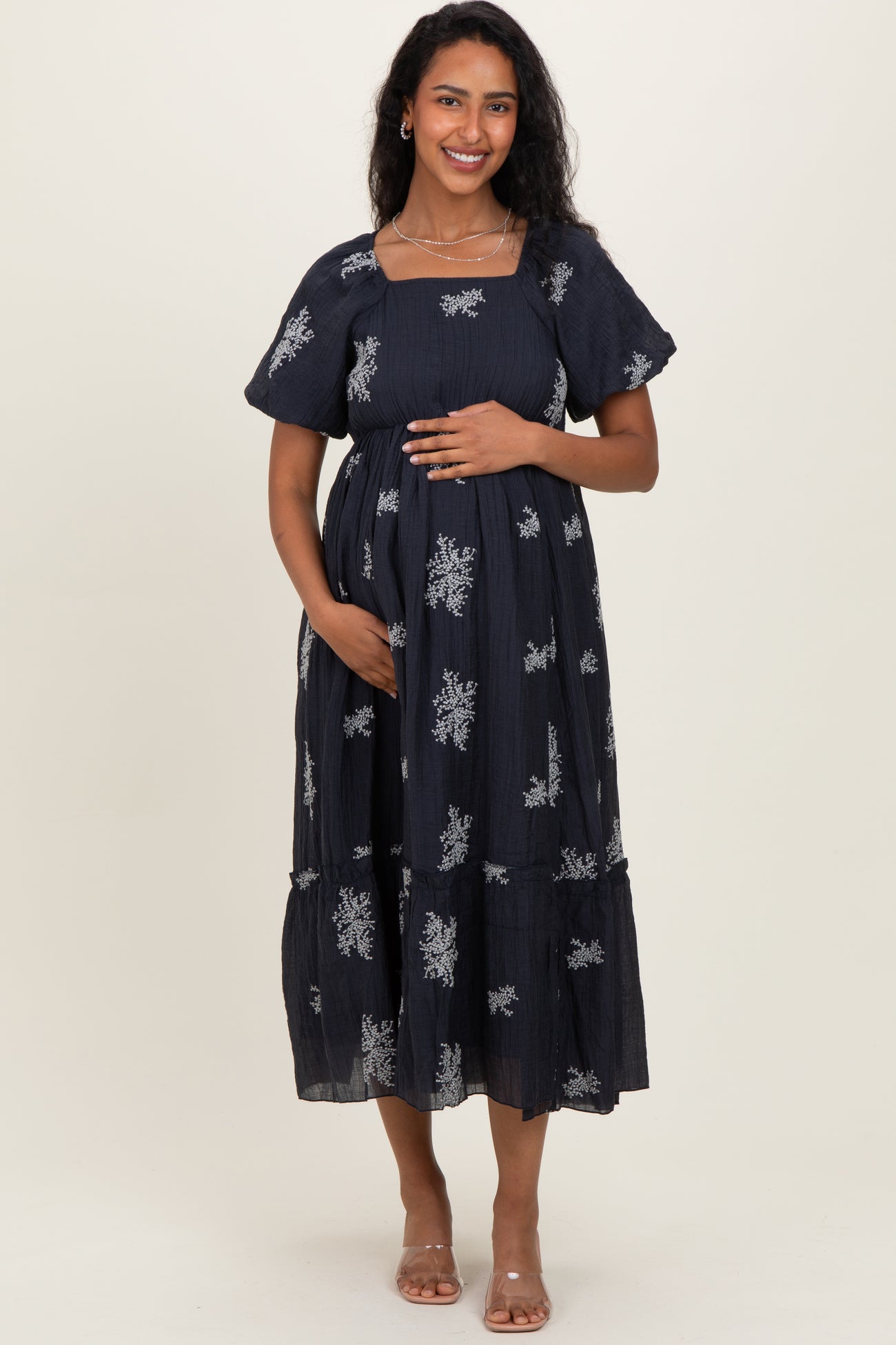 woman wearing a navy blue maternity dress with silver floral patterns and a baby bump, standing and smiling