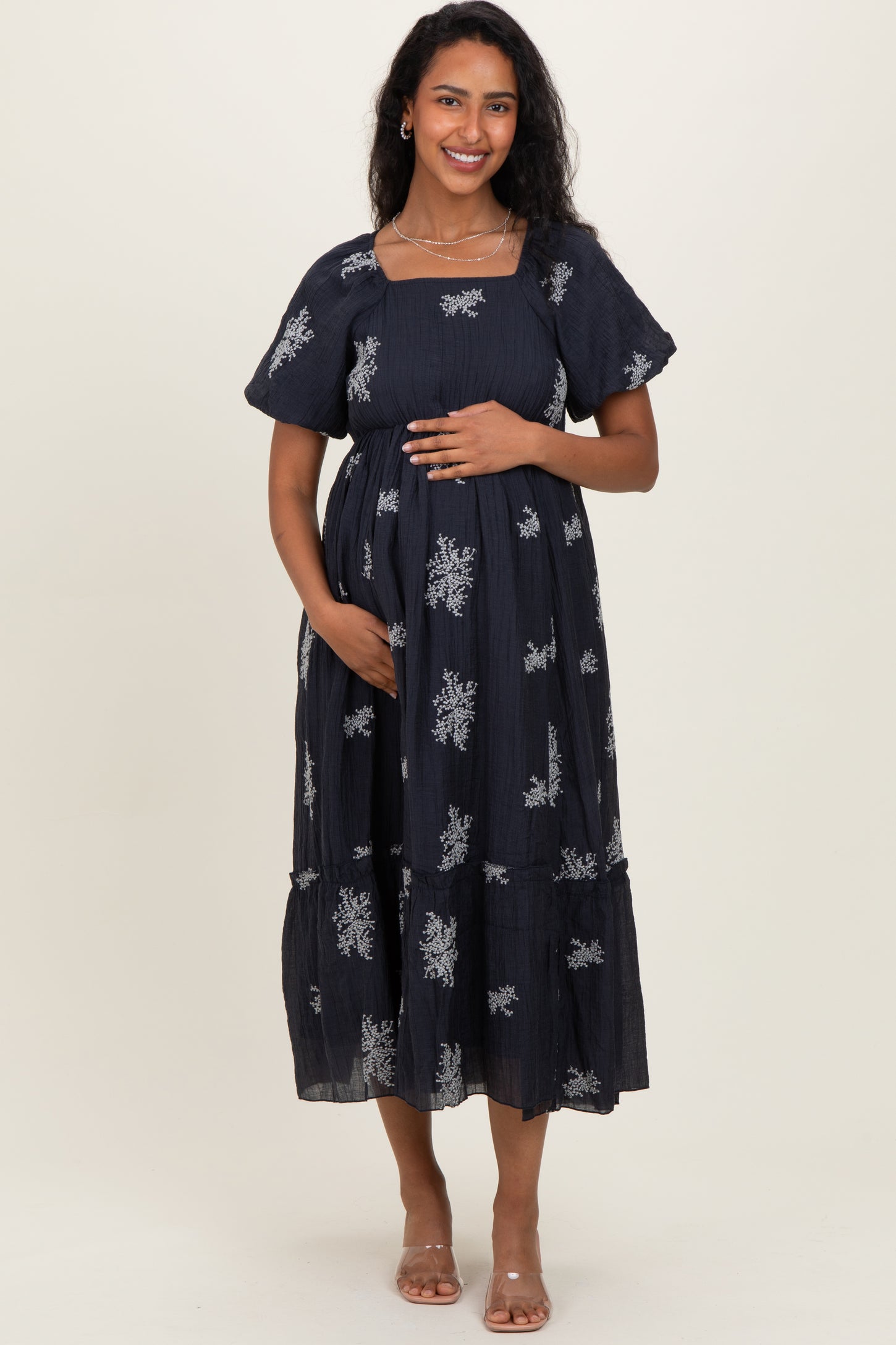 woman wearing a navy blue maternity dress with silver floral patterns and a baby bump, standing and smiling