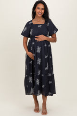 woman wearing a navy blue maternity dress with silver floral patterns and a baby bump, standing and smiling