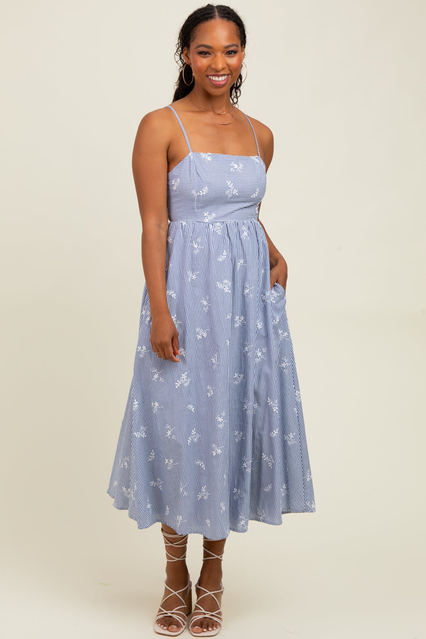 woman wearing a blue striped midi dress with floral patterns and thin straps, standing with hands in pockets