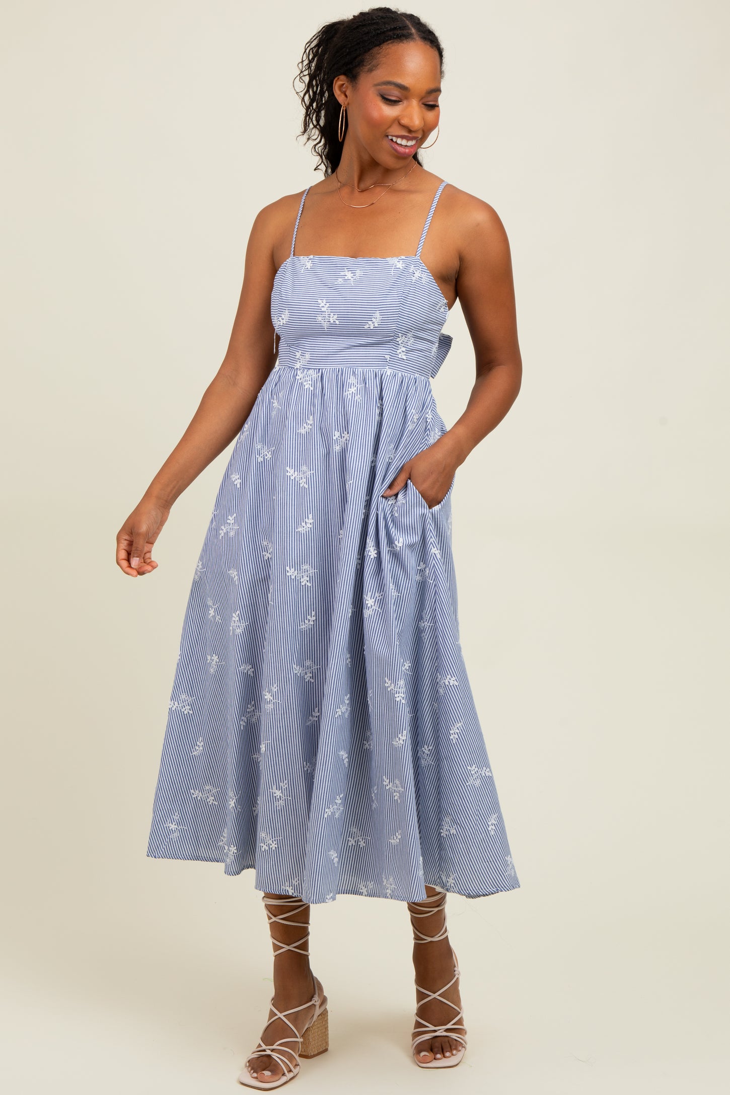 woman wearing a blue and white striped midi dress with floral details and adjustable straps, standing with one hand in a pocket