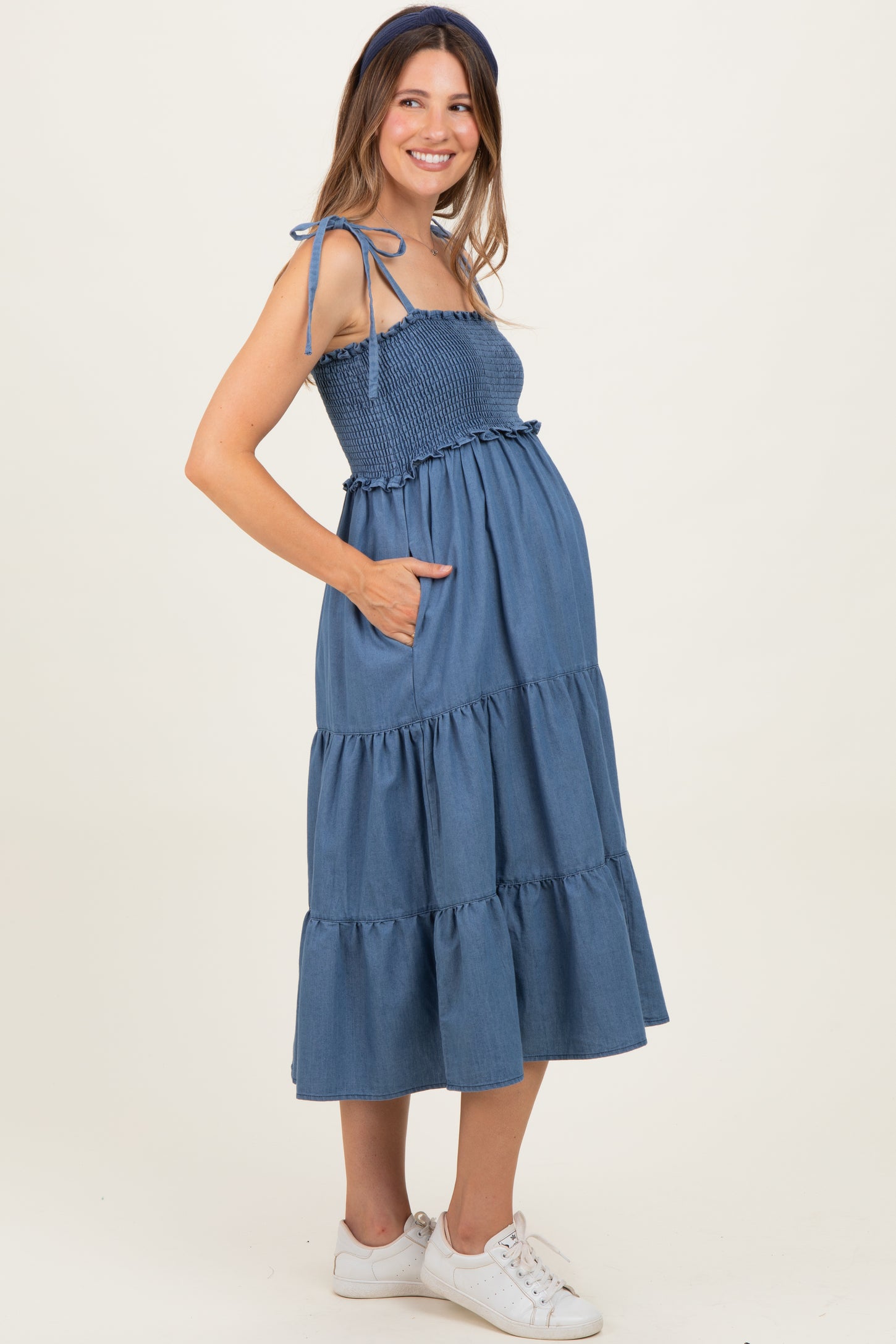 woman wearing a blue tiered maternity dress with adjustable straps and pockets, showing her baby bump