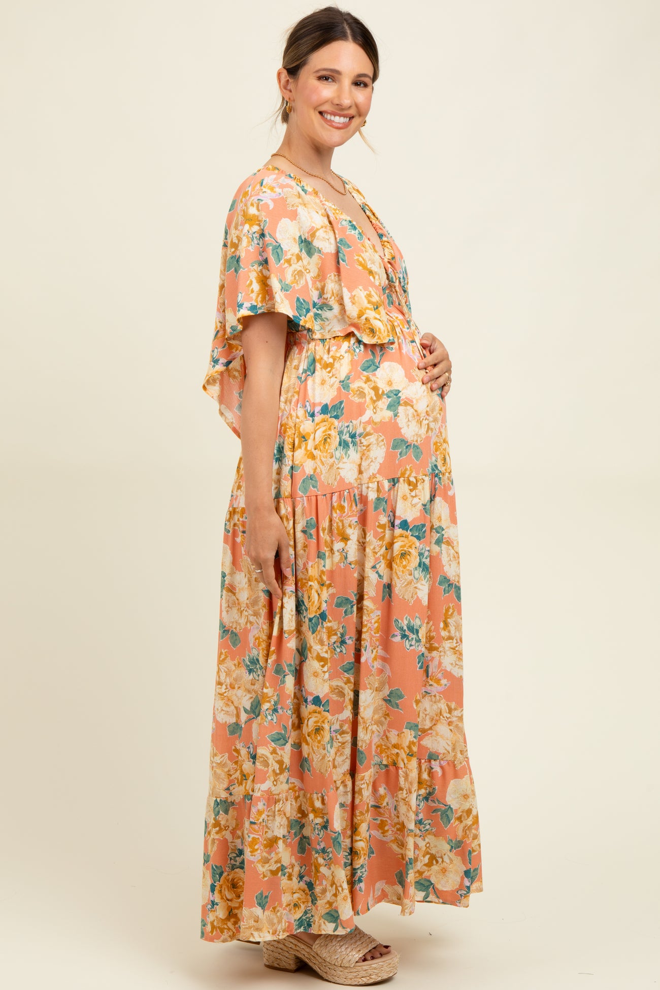 woman wearing a floral maternity maxi dress in peach tones, holding her baby bump, styled with a flowy cape detail