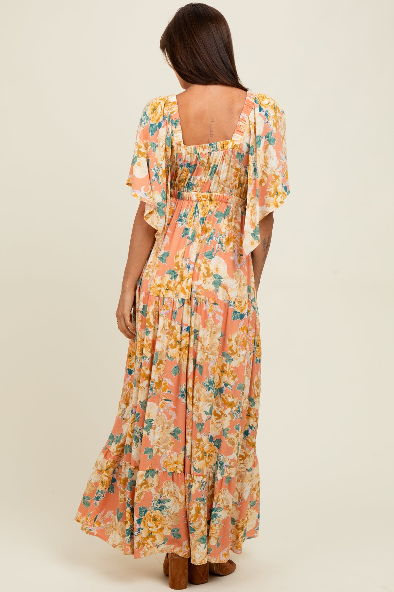 woman wearing a floral maxi dress with flutter sleeves and a fitted bodice in peach and yellow tones, viewed from the back