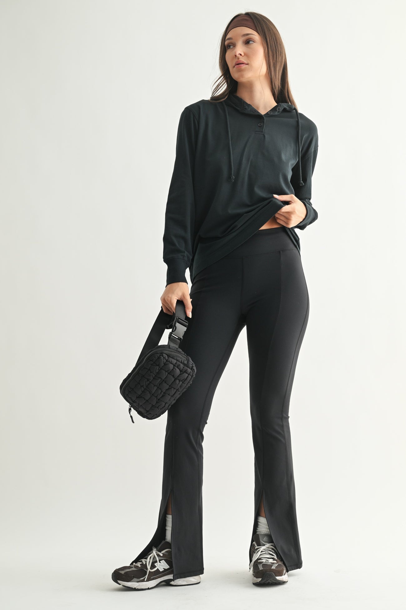 woman wearing a black long sleeve hoodie and black flared leggings with side slits, holding a small black bag