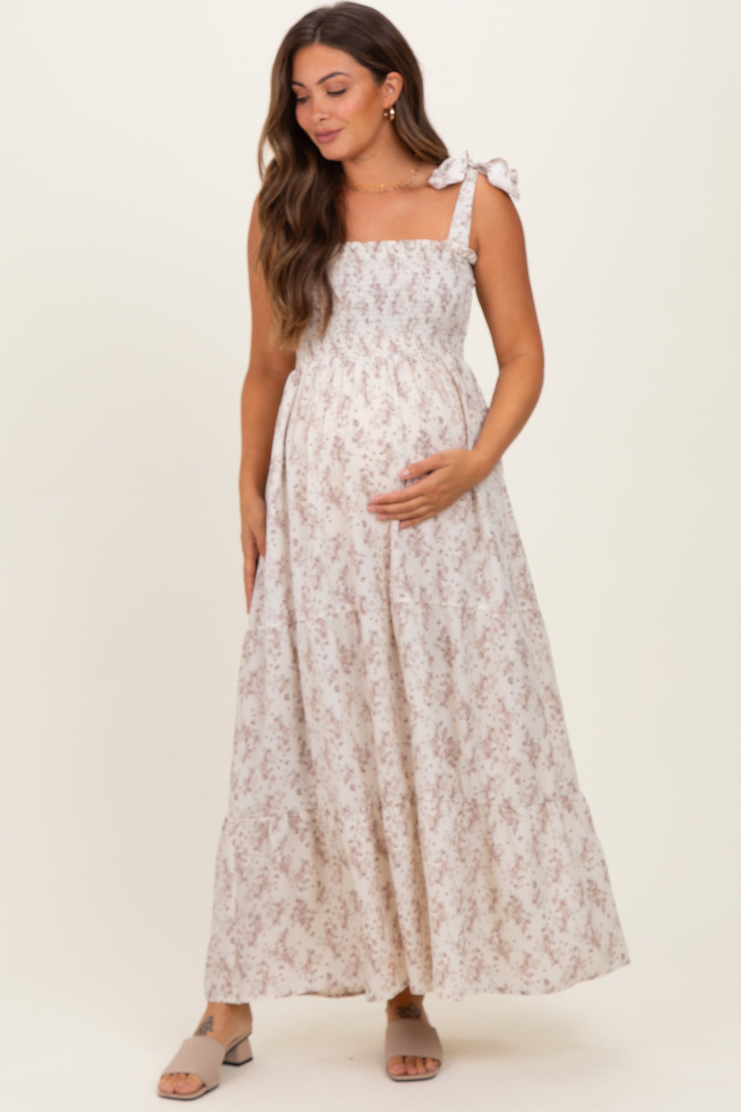 woman wearing a maternity maxi dress with floral print and bow straps, gently cradling her baby bump
