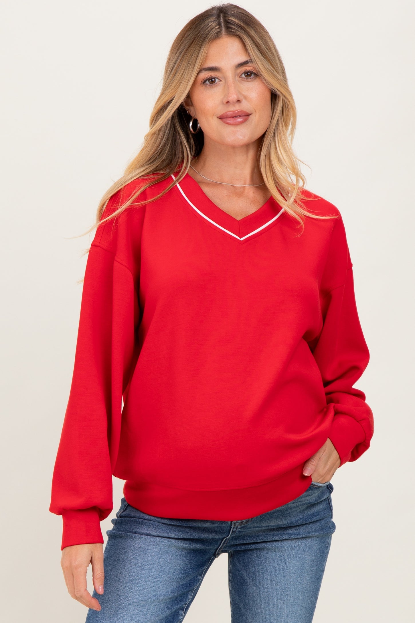 woman wearing a red maternity sweatshirt with a v-neck and white trim, paired with blue jeans and a baby bump visible