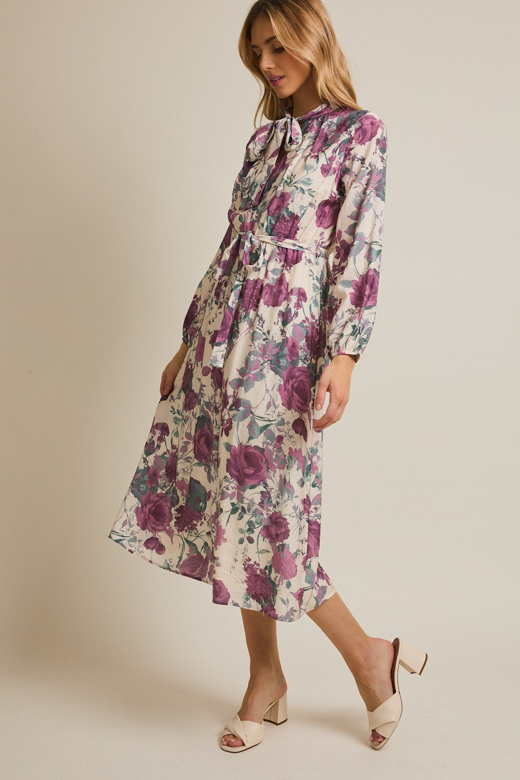 woman wearing a floral long sleeve midi dress with a tie neck and belted waist, standing on an angle