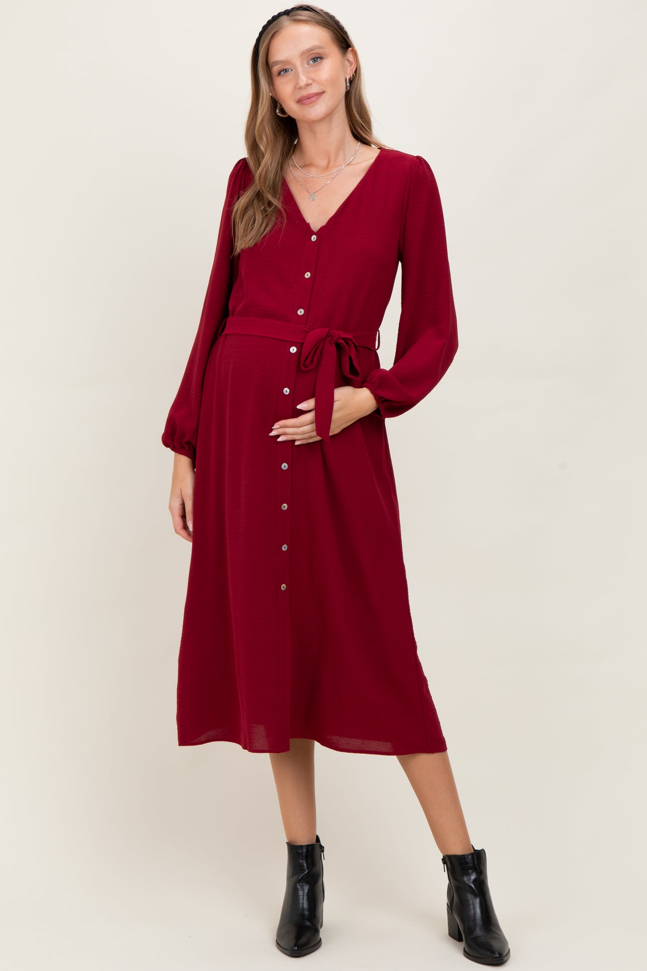 woman wearing a burgundy maternity dress with long sleeves, button details, and a tie waist showcasing her baby bump