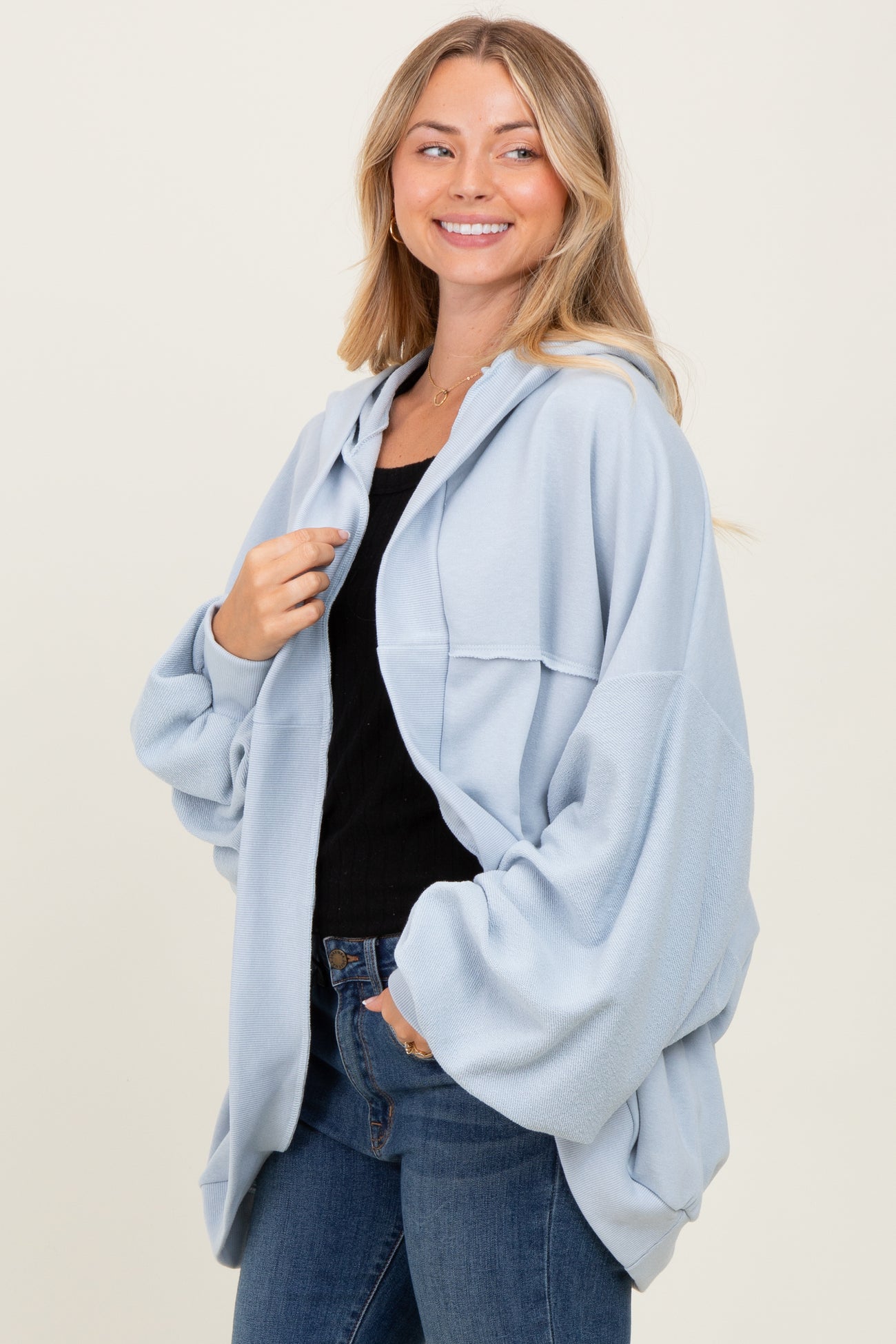 woman wearing a light blue oversized hoodie with a front pocket and black top underneath, smiling against a neutral background