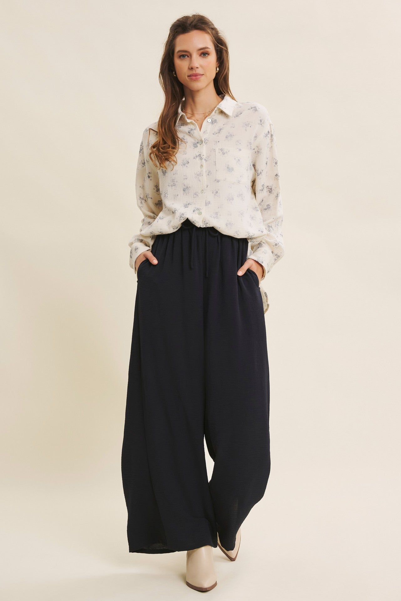 woman wearing a white floral long sleeve button-up shirt and navy wide-leg pants, standing and posing