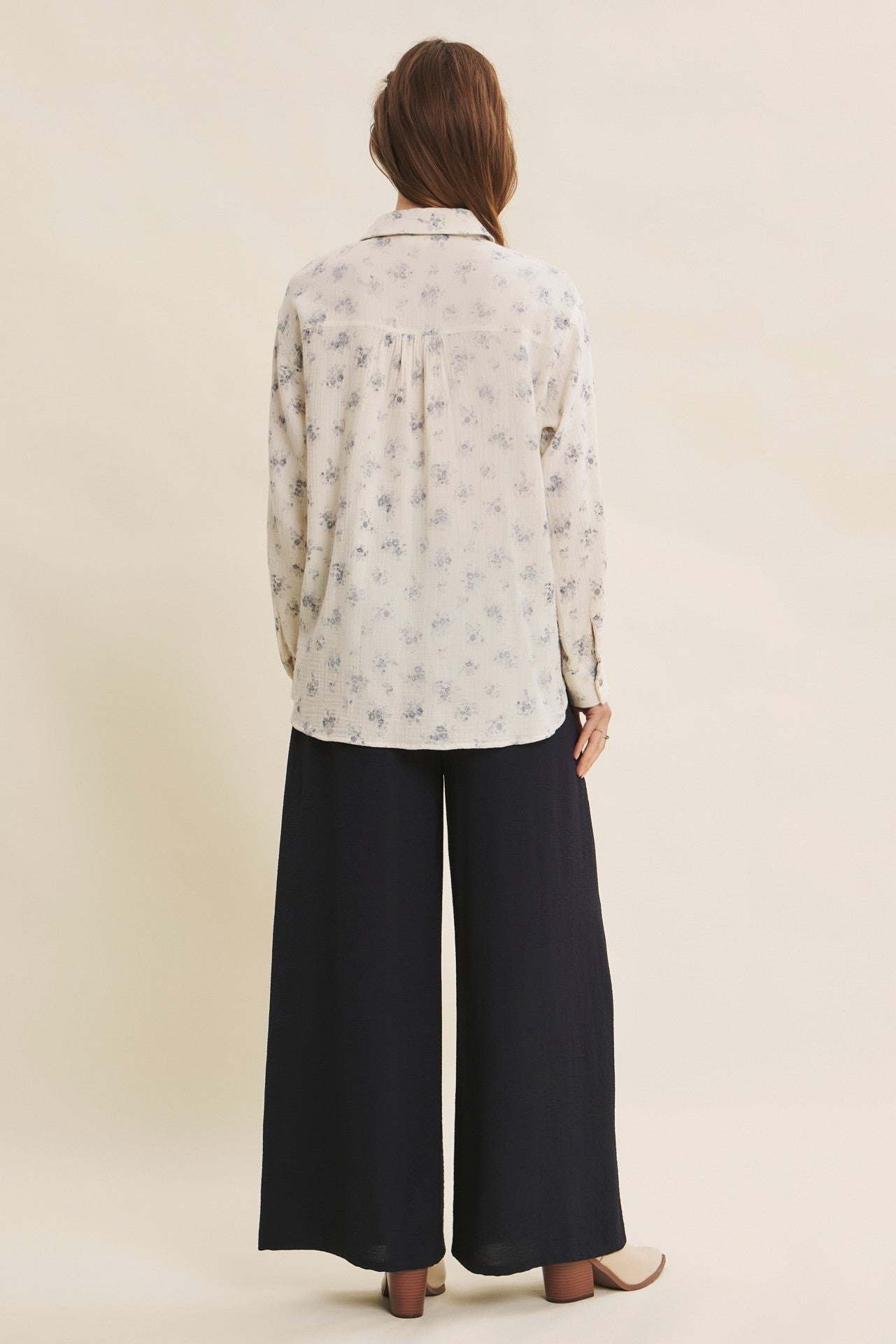 woman wearing a white long sleeve blouse with a floral print and dark wide-leg pants, viewed from the back