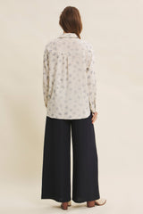 woman wearing a white long sleeve blouse with a floral print and dark wide-leg pants, viewed from the back