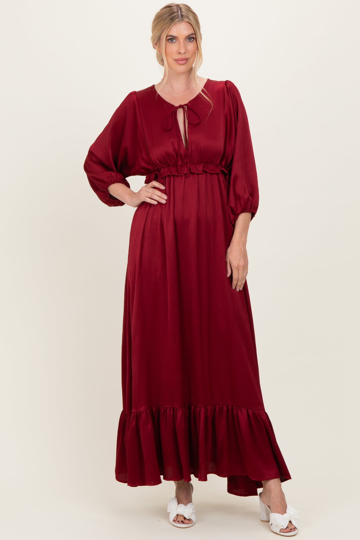 woman wearing a burgundy maxi dress with puff sleeves and a ruffled hem, styled with a tie neckline and white sandals