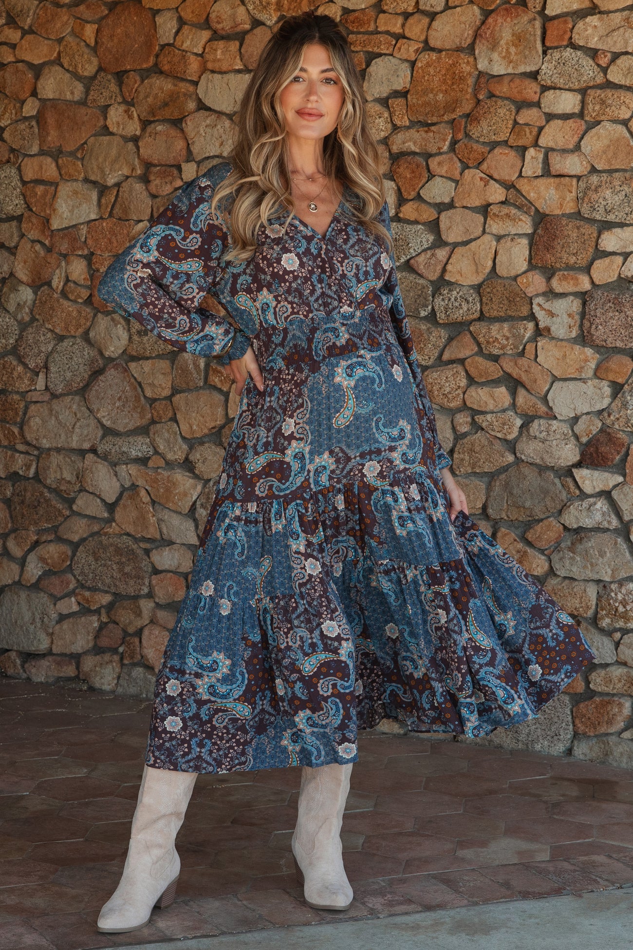 woman wearing a paisley print maternity maxi dress with long sleeves and tiered skirt, posing in front of a stone wall