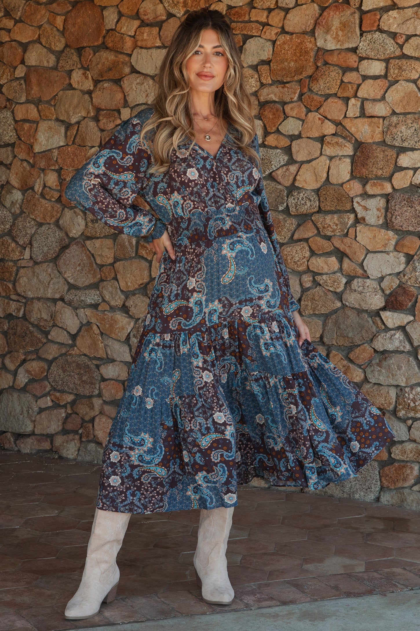 woman wearing a paisley print maternity maxi dress with long sleeves and tiered skirt, posing in front of a stone wall