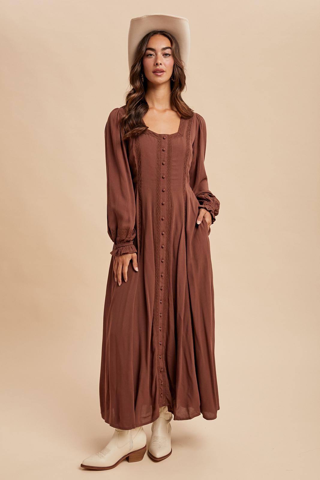woman wearing a long brown button-up dress with puff sleeves and a wide-brimmed hat, standing against a neutral background