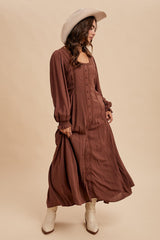 woman wearing a brown long sleeve maxi dress with buttons down the front and a wide-brimmed hat, standing in a flowing pose