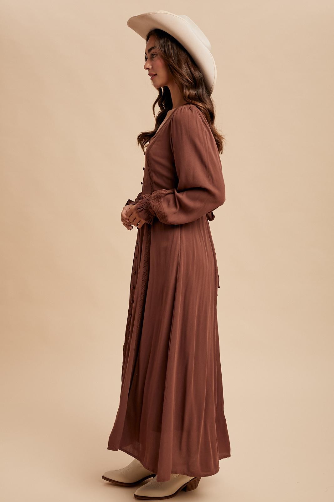 woman wearing a brown long sleeve maxi dress with button details and a hat, styled in a side profile view
